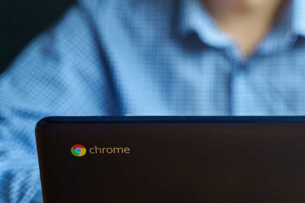 Chrome logo on lid of Chromebook laptop while man in blue shirt uses it