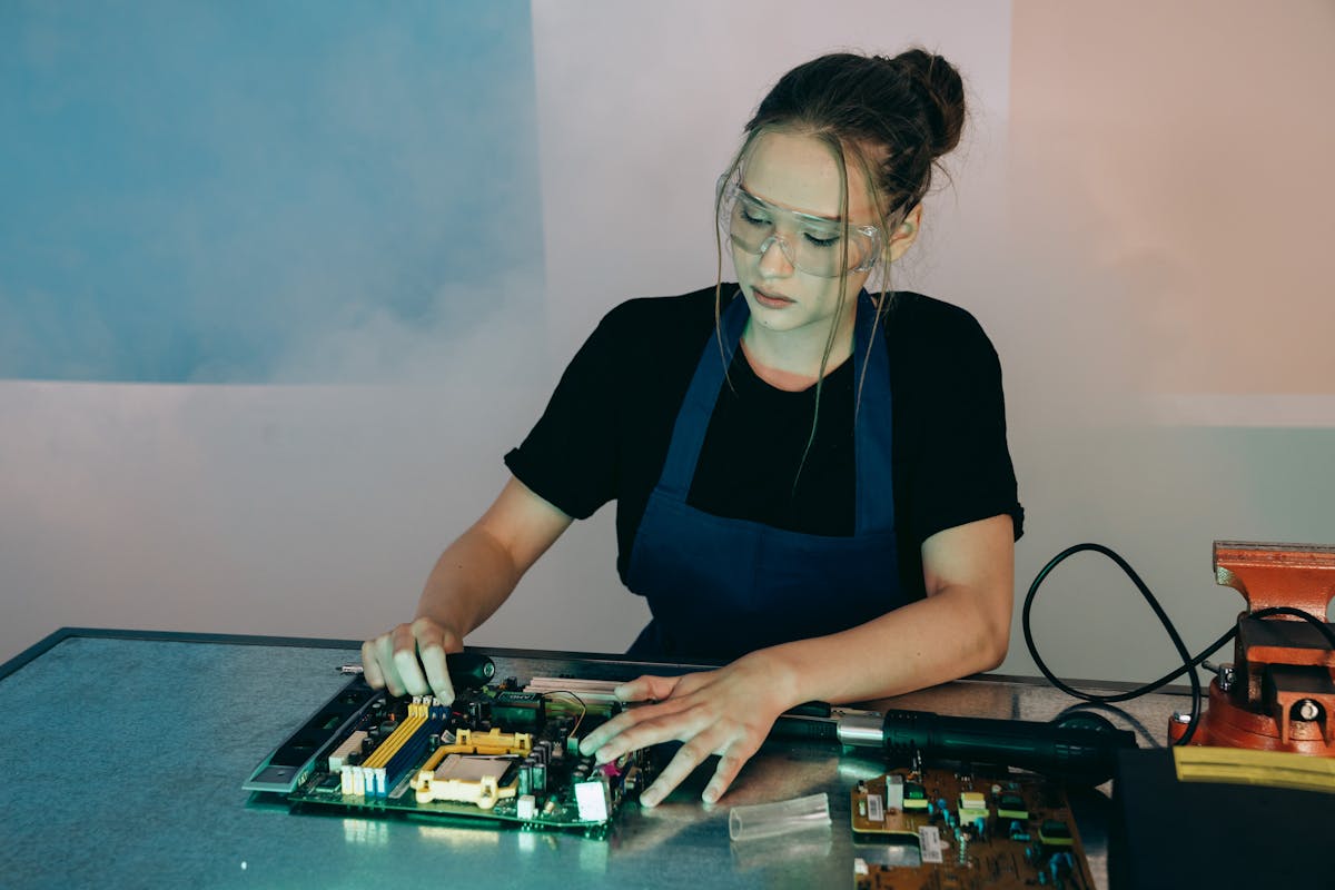 Young woman in protective goggles soldering electronics on a workbench indoors.