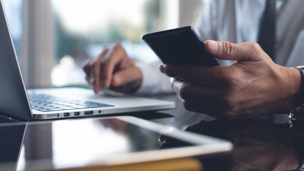 Man holding phone while using laptop and tablet on a desk