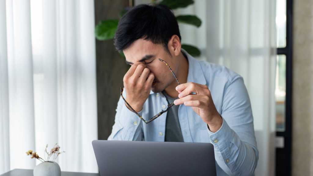 Man pinching tired eyes with glasses off while working at laptop on desk
